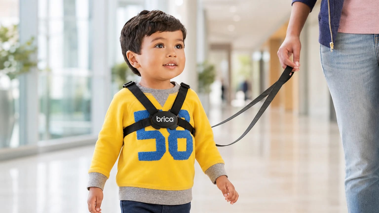 A toddler wears black Brica walking reins while a nearby adult loosely holds the lead in a bright modern indoor space.