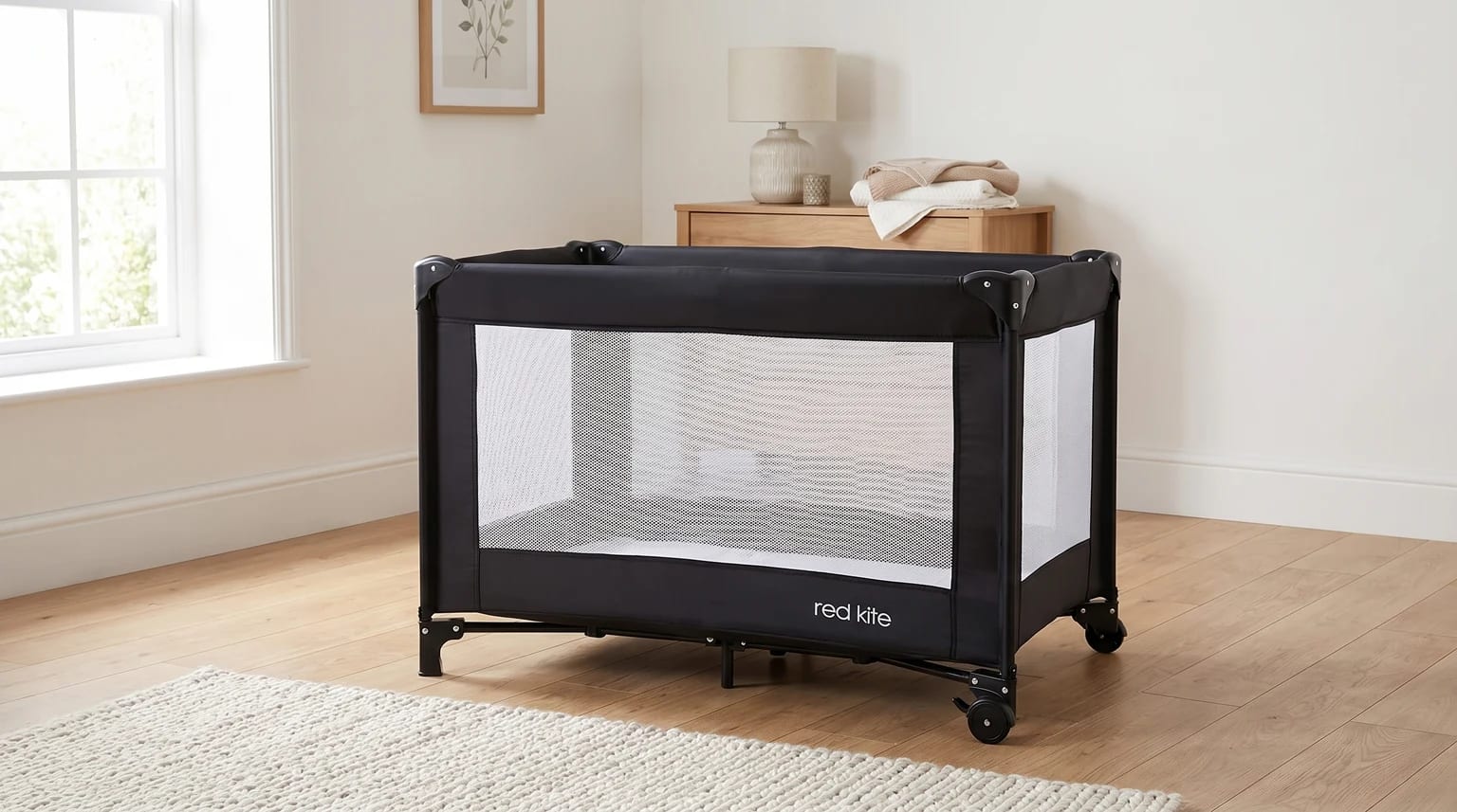 A black Red Kite Sleeptight travel cot with white mesh sides set up empty in a bright room.
