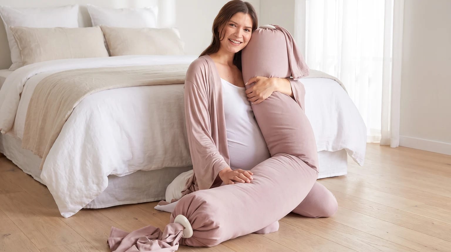 A pregnant woman sits on a wooden floor in a bright bedroom, smiling as she hugs a long, dusty pink pregnancy pillow.