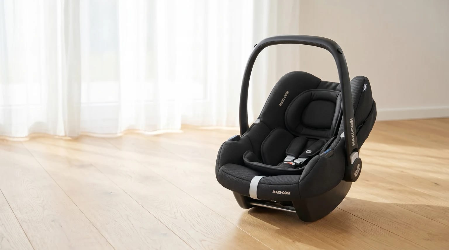 A black Maxi-Cosi infant carrier sitting on a light wooden floor in a brightly lit room with sheer curtains in the background.