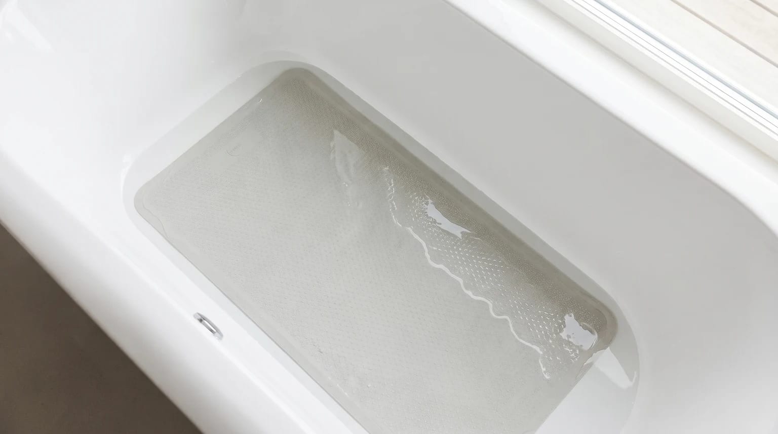 A light grey, textured bath mat lying at the bottom of a clean white bath, with a shallow layer of water over it.