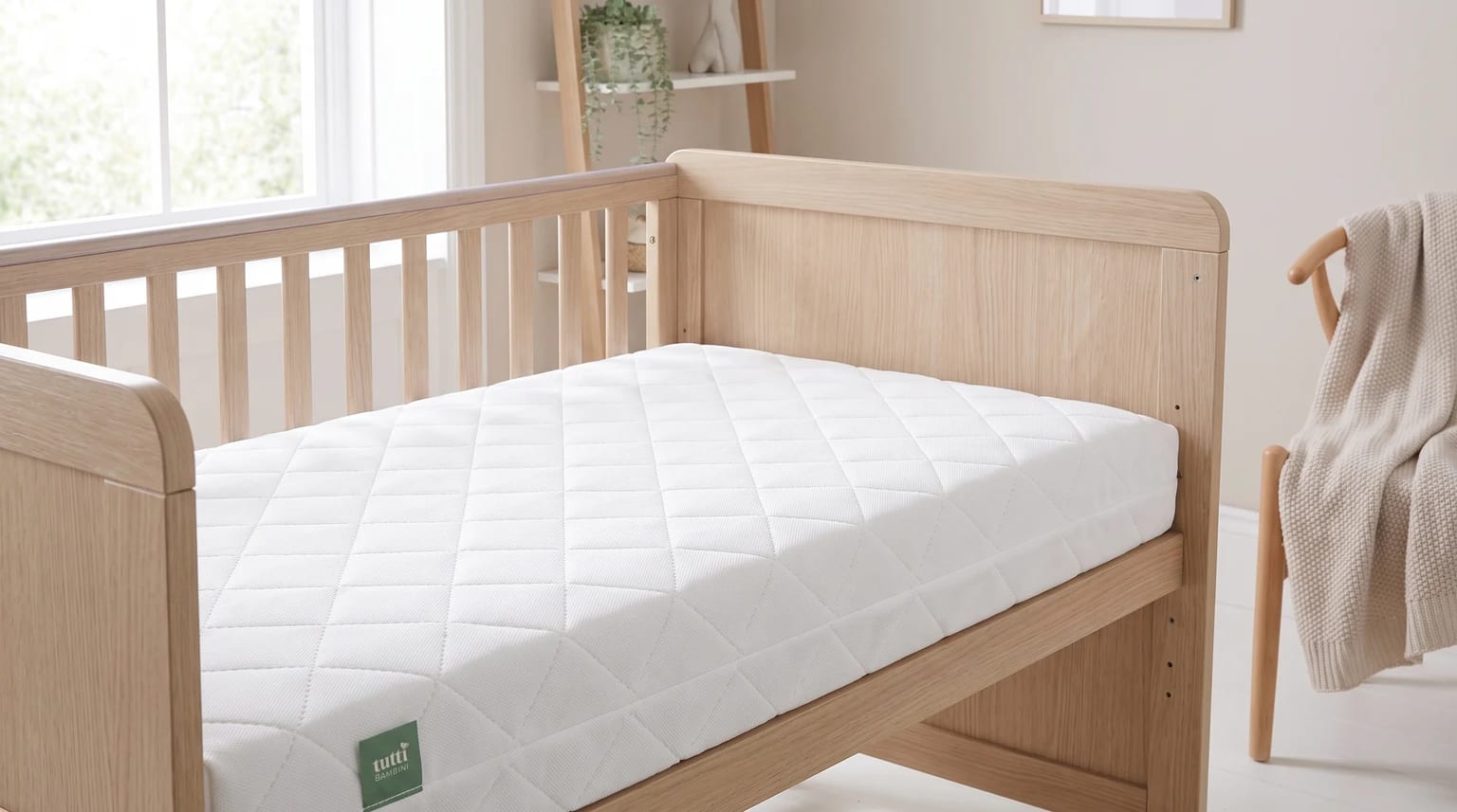 A white cot bed mattress with a diamond-quilted cover, placed inside a light oak cot bed in a neutral-toned nursery.
