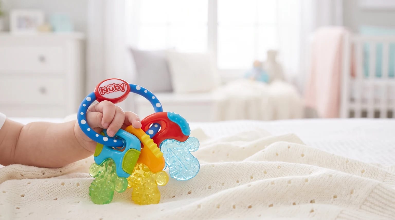 A baby's hand holds a colourful Nuby key-ring teether on a cream knitted blanket, with a nursery cot in the background.