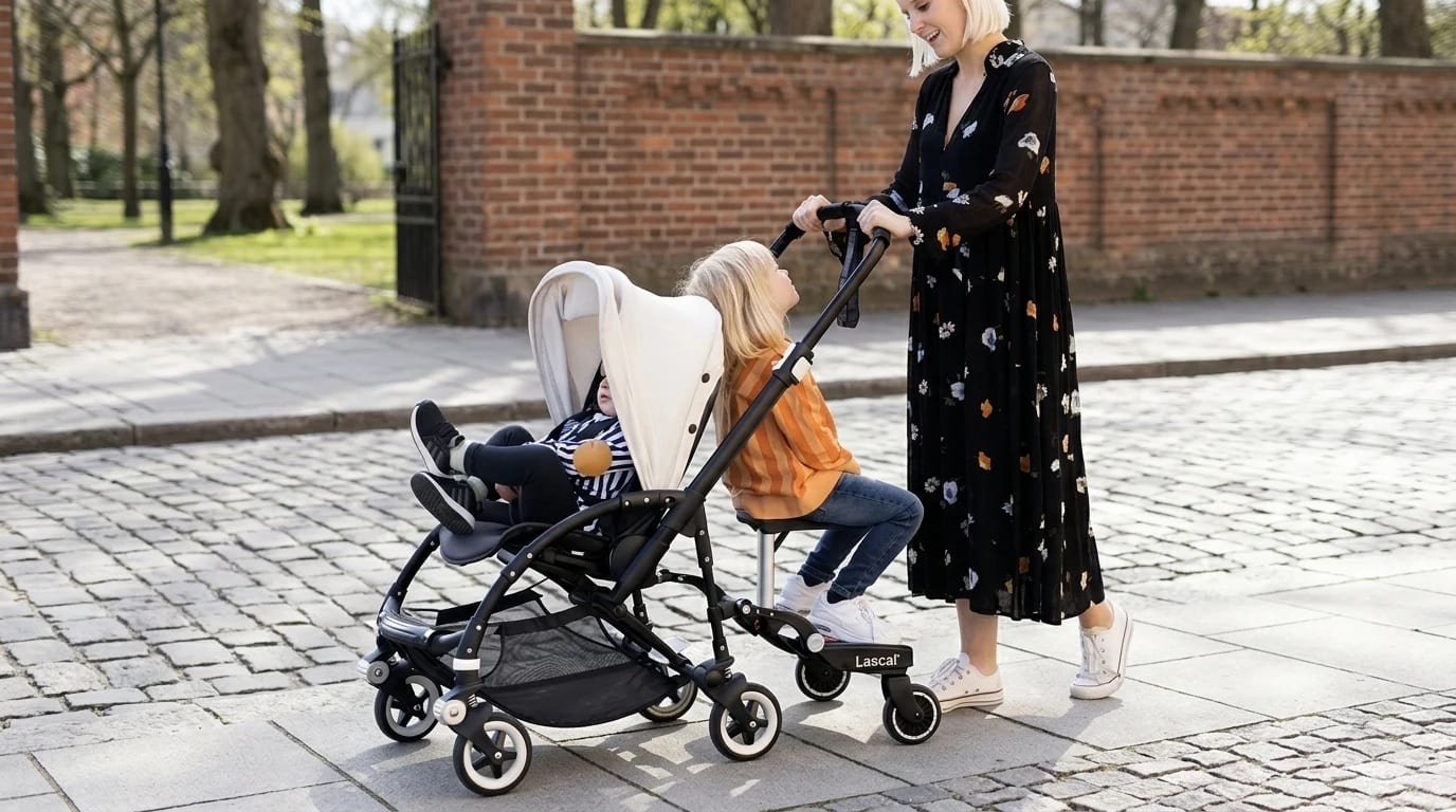 A child sits on a saddle-seat buggy board attached behind a grey pushchair on a sunny pavement.
