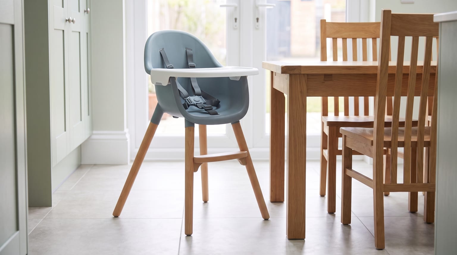 An empty blue-grey high chair with wooden legs stands on a tiled floor next to a wooden dining table in a bright kitchen-diner.