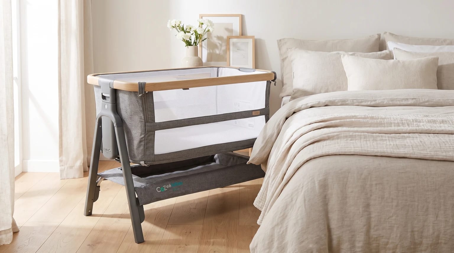 A grey bedside crib with a wood-effect trim and mesh sides, set up in a calm bedroom next to a bed with neutral linen.