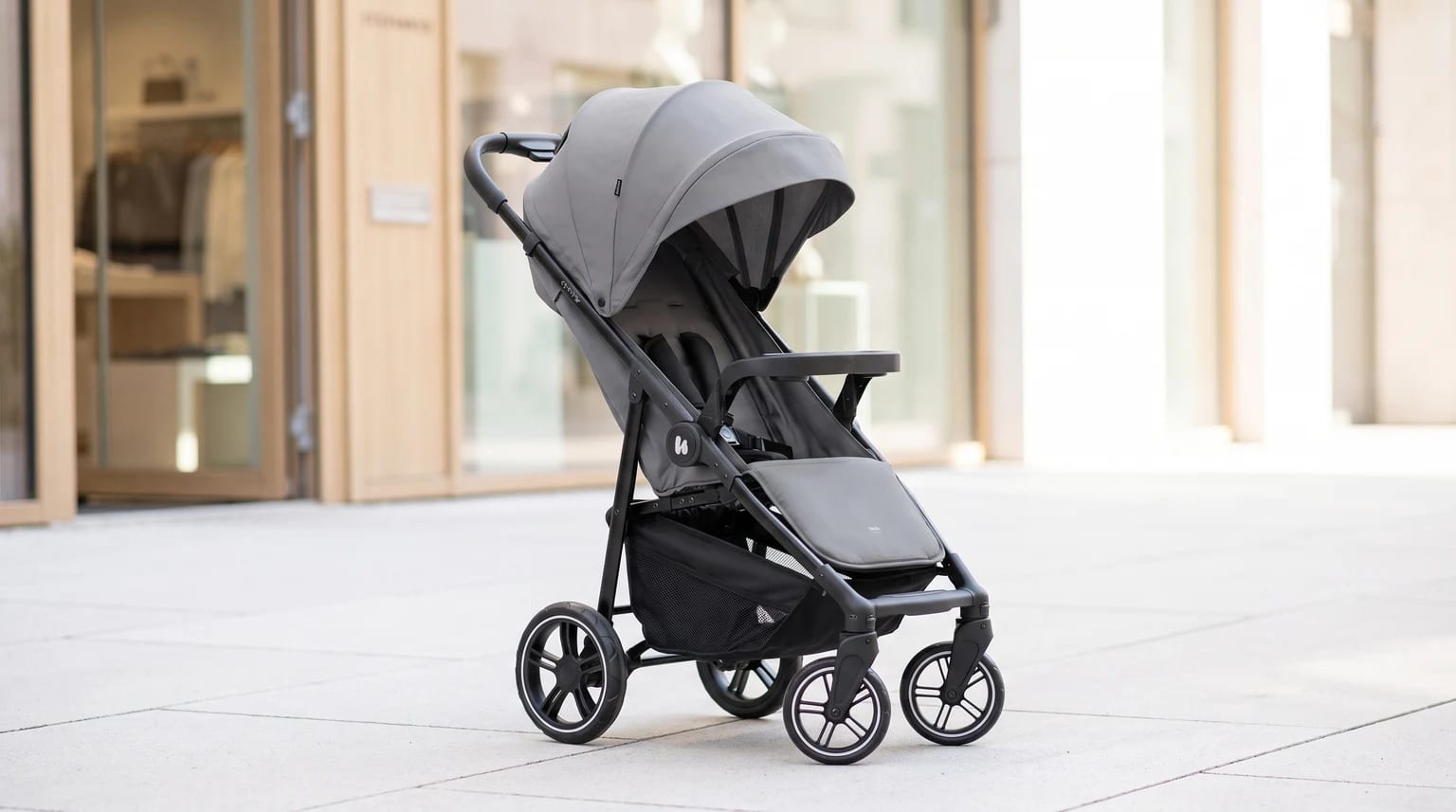 A modern grey pushchair with a black frame and large sun canopy, pictured on a pavement in front of a shop.