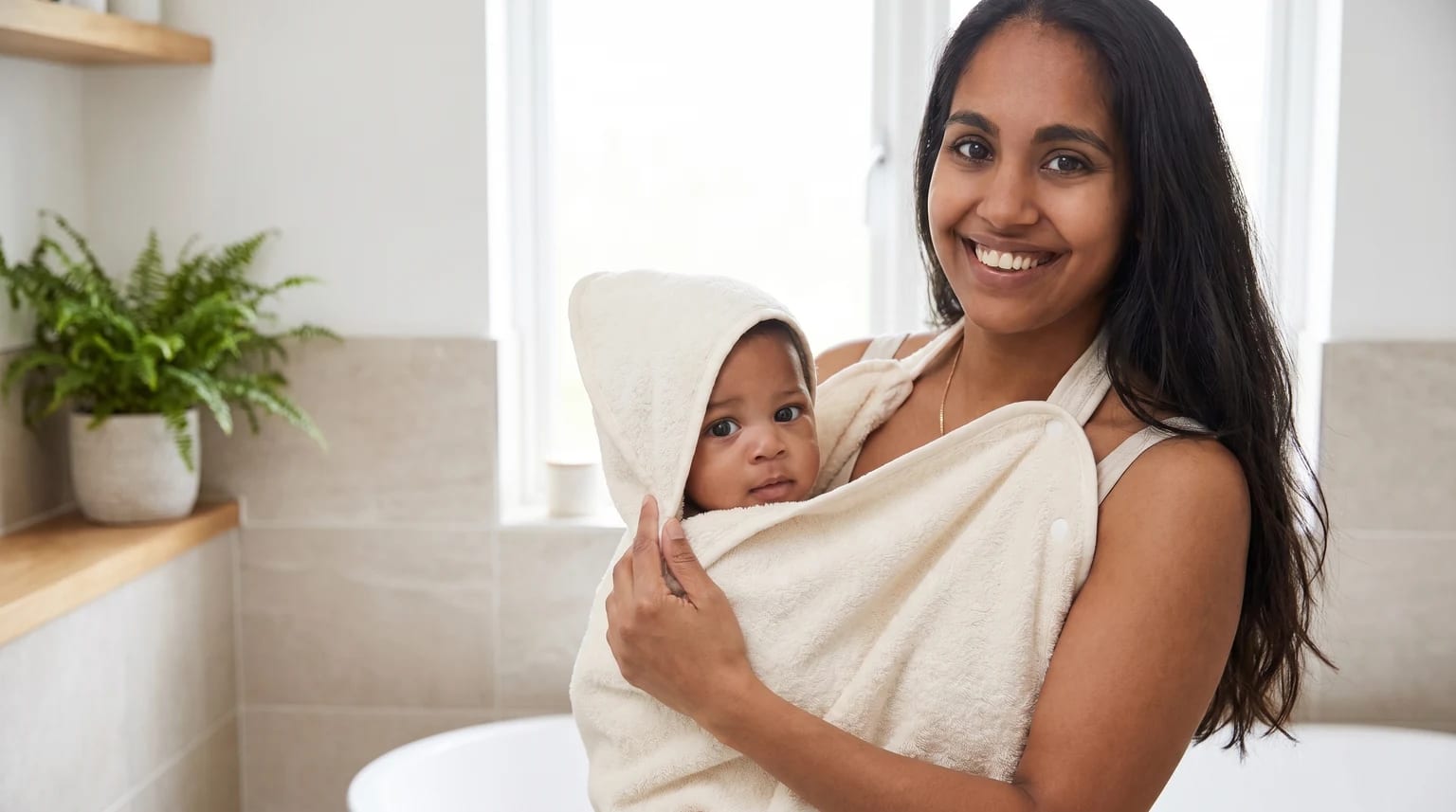 A smiling mother holds her baby, who is wrapped snugly in a cream-coloured hooded towel, in a modern bathroom.