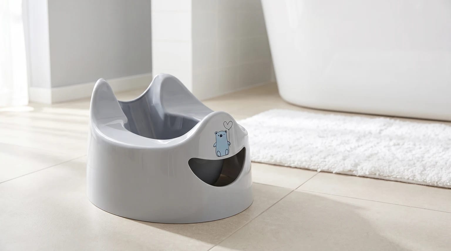 A light grey potty with a cartoon blue bear holding a heart balloon, placed on a tiled bathroom floor.