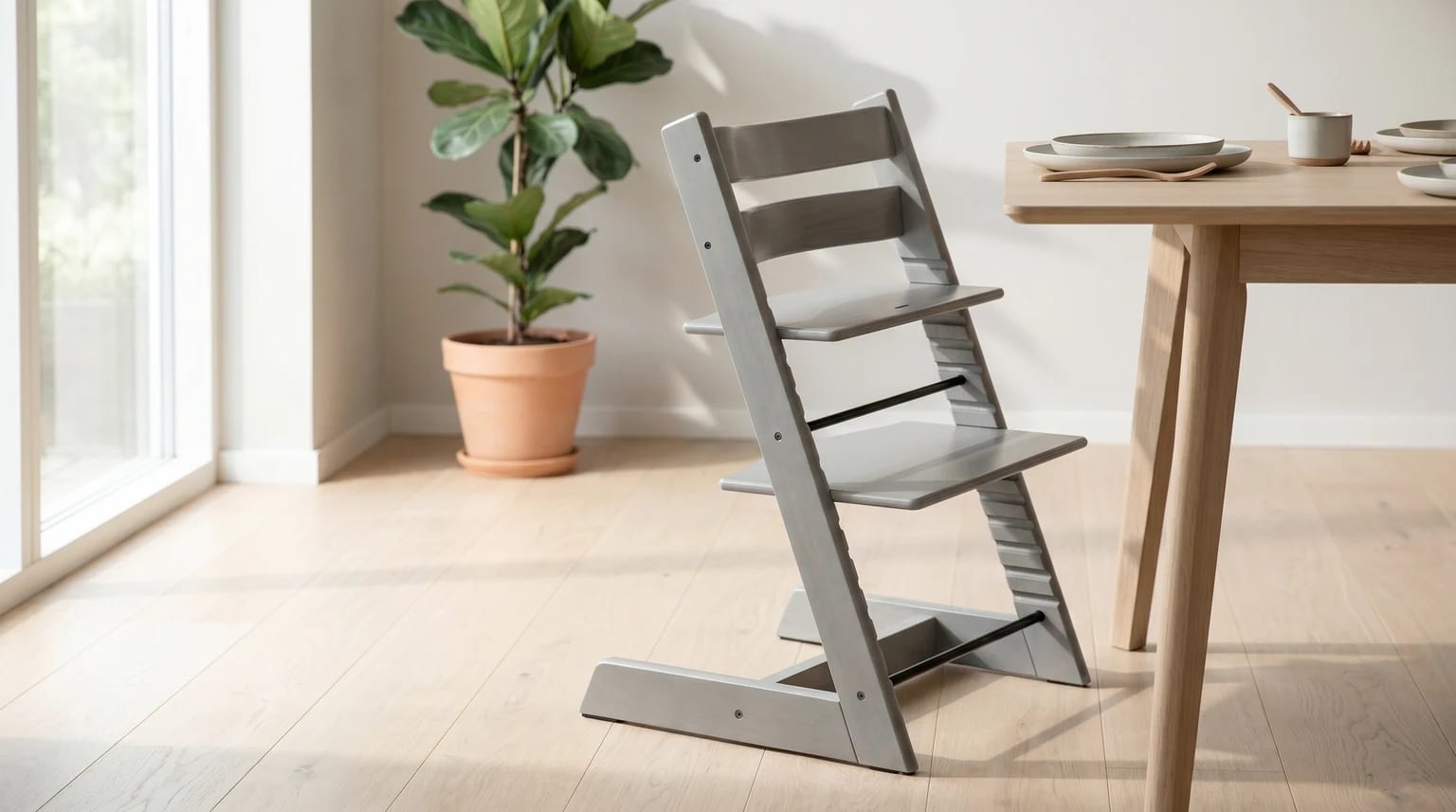 A grey wooden high chair pulled up to a light wood dining table in a brightly lit room with a large houseplant.