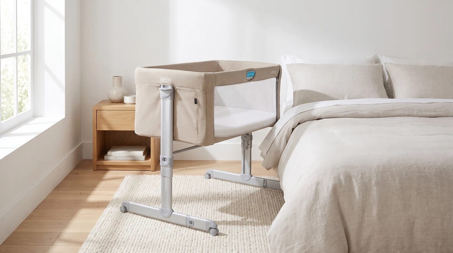 A beige bedside crib with a mesh side panel, positioned on a textured rug next to an adult's bed in a sunlit bedroom.