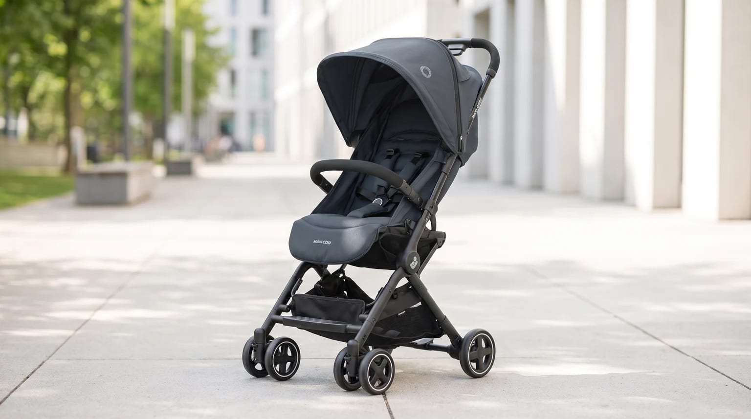A charcoal grey pushchair stands empty on a sunny, paved walkway in a modern city setting.
