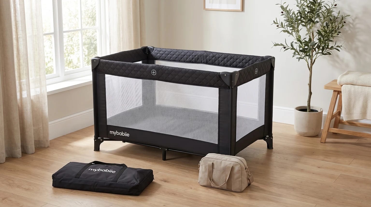 A black My Babiie travel cot with quilted trim set up empty in a sunlit room, with its carry bag nearby.