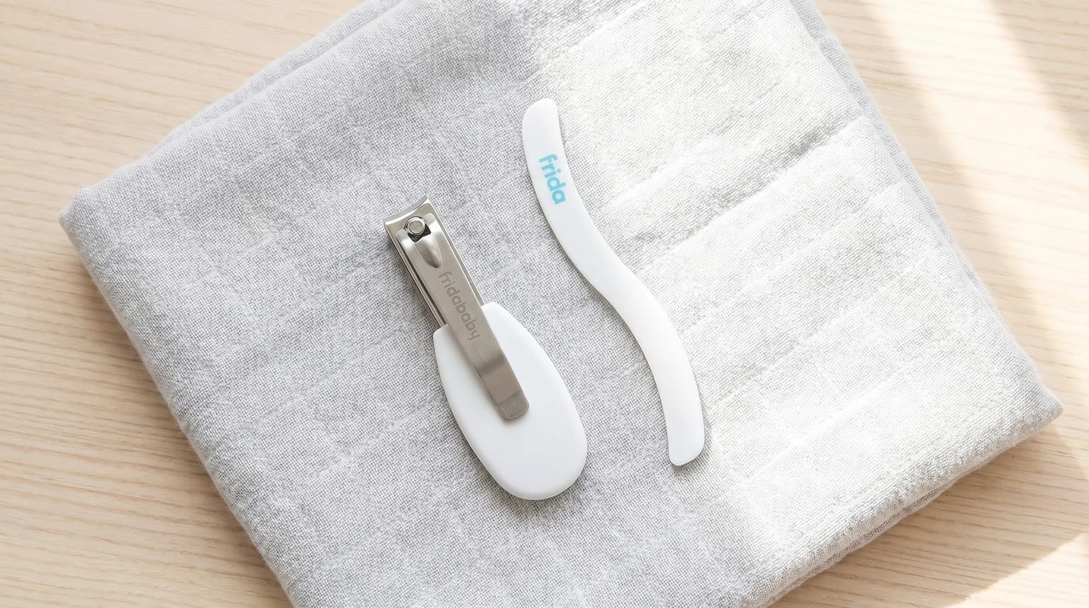 A pair of white Fridababy nail clippers and a matching S-shaped nail file resting on a folded grey muslin cloth.